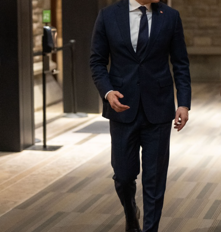 Man in a navy suit walking down a hallway, smiling, in an indoor office-like setting with brick walls.