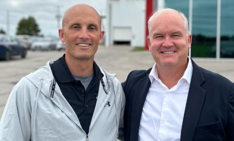 Two smiling men stand side by side outdoors, one in a light jacket and the other in a suit jacket, posing for a photo in front of a modern building.