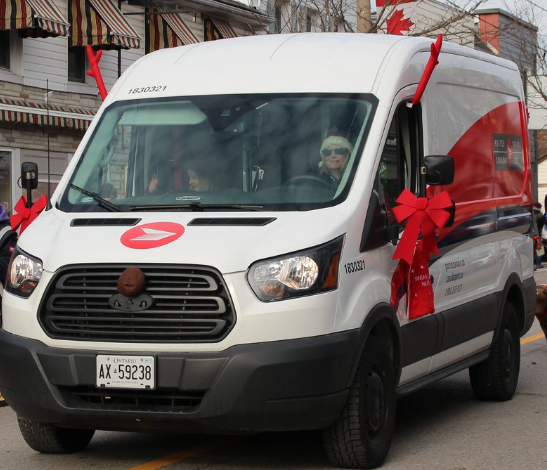 White cargo van in a street parade decorated with large red bows on its mirrors and front grille.