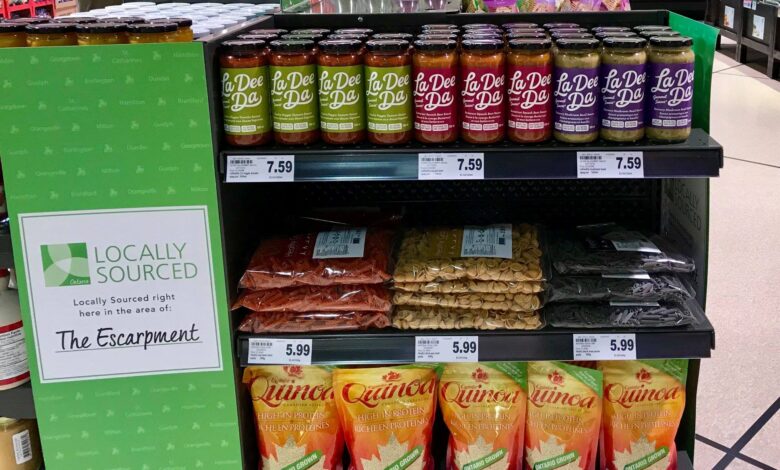 Shelf display of La Dee Da sauces in green and red jars with Ontario-grown quinoa bags below and a green Locally Sourced sign on the left.