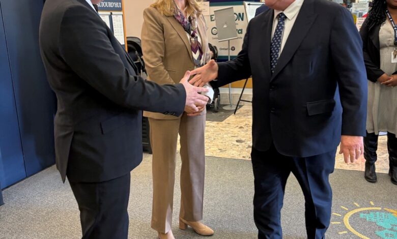 Two men in suits shake hands in a bright lobby while a smiling woman in a tan suit stands between them near an orange wall with 'QUEENSWAY' signage.