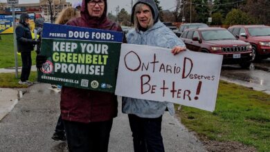 Two people in rain gear stand on a wet street holding protest signs; one sign reads 'KEEP YOUR GREENBELT PROMISE!' and the other says 'Ontario Deserves Better!'