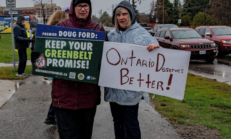 Two people in rain gear stand on a wet street holding protest signs; one sign reads 'KEEP YOUR GREENBELT PROMISE!' and the other says 'Ontario Deserves Better!'