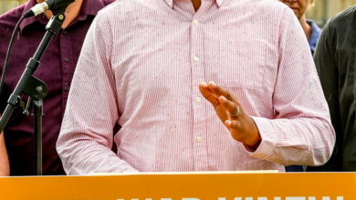 Wab Kinew speaks at a podium with an orange banner reading 'WAB KINEW FOR ALL OF US'; supporters stand behind him.