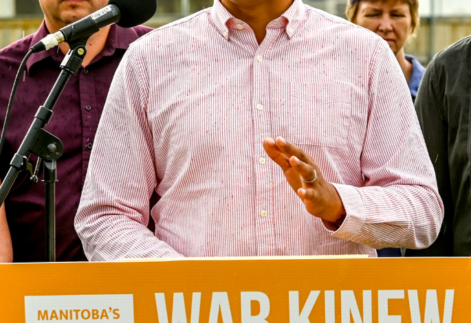 Wab Kinew speaks at a podium with an orange banner reading 'WAB KINEW FOR ALL OF US'; supporters stand behind him.