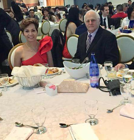 Woman in a red formal dress and a man in a suit sit at a banquet table during a formal event; table is set with bowls, bread, and a blue water bottle.