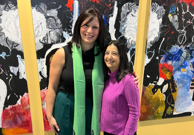 Two women smiling and posing together in front of a vibrant abstract mural.
