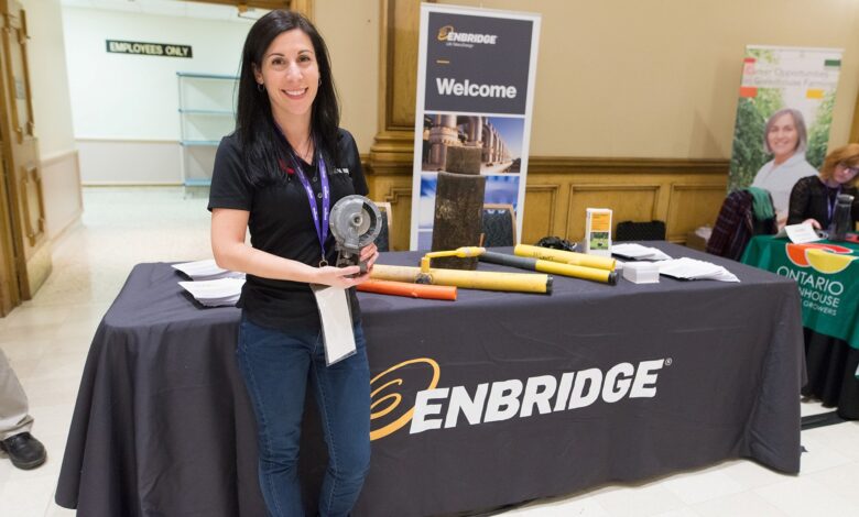 Woman at an ENBRIDGE booth holding a pipe tool, standing behind a table with promotional materials.