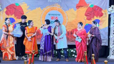 Group of people in colorful sarees on a stage during a cultural event; one woman speaks into a microphone while others look on.