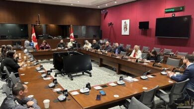 People seated around a U-shaped conference table in a formal meeting room with Canadian flags and microphones on each seat.