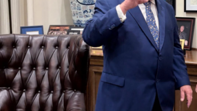 Man in a blue suit gesturing beside a wooden desk in an office, with a large blue‑and‑white vase and framed photos behind him.
