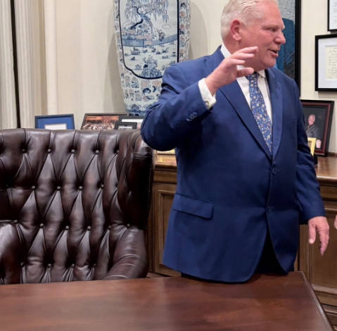 Man in a blue suit gesturing beside a wooden desk in an office, with a large blue‑and‑white vase and framed photos behind him.