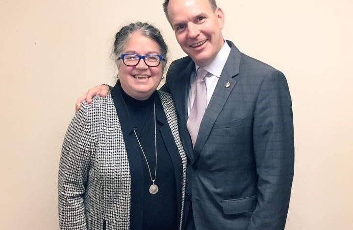 Two coworkers stand side by side and smile for the camera, the man in a gray suit and light tie and the woman in a checkered blazer, black top, and red skirt with her arm around his back by a beige wall.