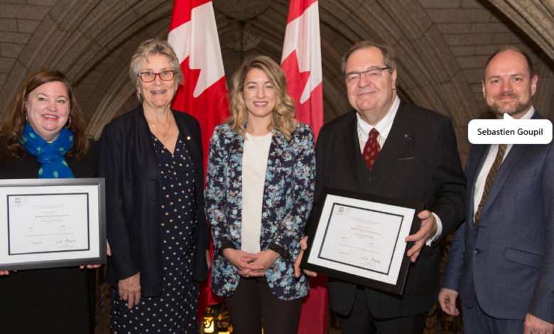 Five professionals pose for a formal ceremony photo; two are holding framed certificates with Canadian flags in the background.