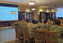 Soldiers in camouflage seated in a conference room watching a presentation on a projector screen at the front.