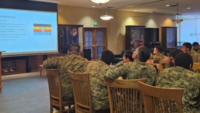 Soldiers in camouflage seated in a conference room watching a presentation on a projector screen at the front.