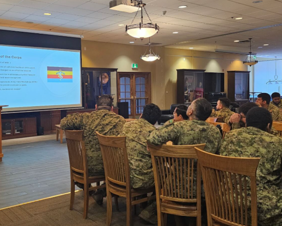 Soldiers in camouflage seated in a conference room watching a presentation on a projector screen at the front.