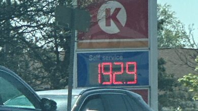Esso gas station sign with blue oval logo and a digital price board displaying 192.9 that’s visible in the foreground of cars at a pump lot.