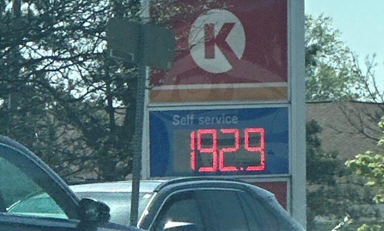 Esso gas station sign with blue oval logo and a digital price board displaying 192.9 that’s visible in the foreground of cars at a pump lot.