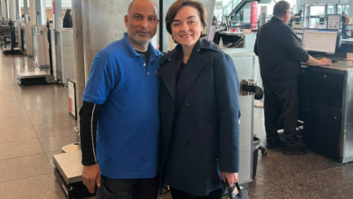 Smiling man in a blue polo shirt and a woman in a dark coat pose together at an airport check-in area near counters and computer terminals.