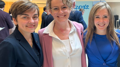 Three smiling women posing together at a professional event, wearing business attire (left in dark suit, center in pink blazer, right in cobalt blue).