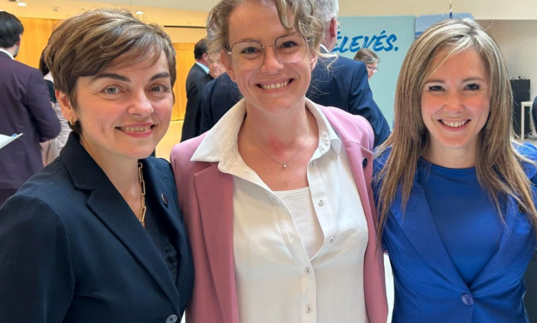 Three smiling women posing together at a professional event, wearing business attire (left in dark suit, center in pink blazer, right in cobalt blue).