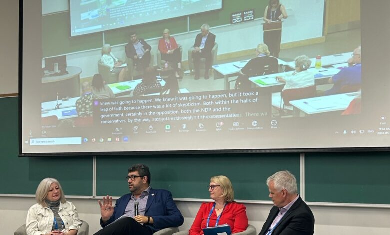 Panel discussion in a classroom: five panelists seated, moderator at a lectern, projected screen behind them showing a live video feed.