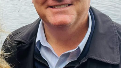 Smiling man with light hair wearing a dark jacket over a blue button-down shirt, standing by a lake with trees in the background.