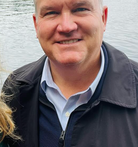 Smiling man with light hair wearing a dark jacket over a blue button-down shirt, standing by a lake with trees in the background.