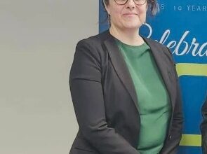 Woman in a green blouse and black blazer standing beside a blue banner, looking at the camera with clasped hands.