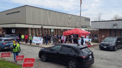 Campaign event outside a brick building with a red canopy, volunteers and voters, and campaign signs along the curb.
