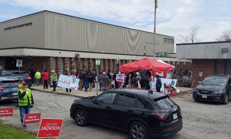 Campaign event outside a brick building with a red canopy, volunteers and voters, and campaign signs along the curb.