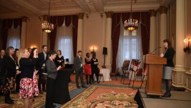 Speaker at a podium addressing a standing audience in an ornate, chandelier-lit ballroom.