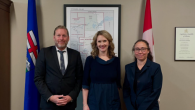 Three professionally dressed adults stand together in a formal office, flanked by flagpoles and a framed map behind them.