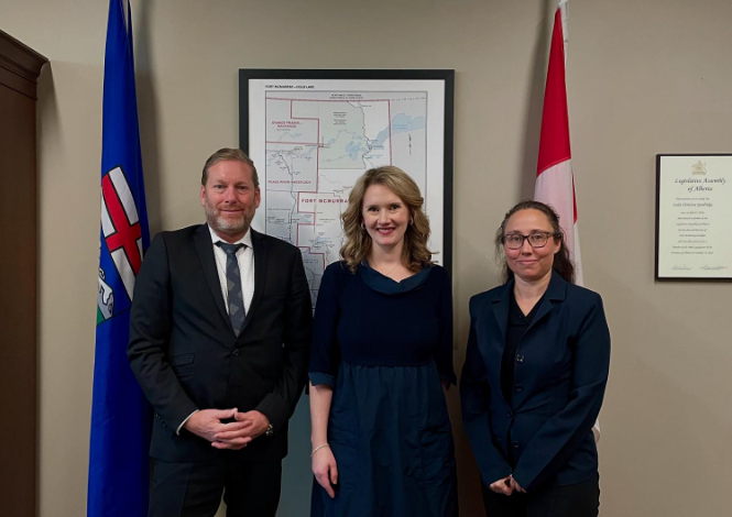 Three professionally dressed adults stand together in a formal office, flanked by flagpoles and a framed map behind them.