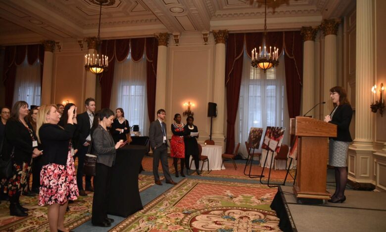 Speaker at a podium addressing a standing audience in an ornate, chandelier-lit ballroom.