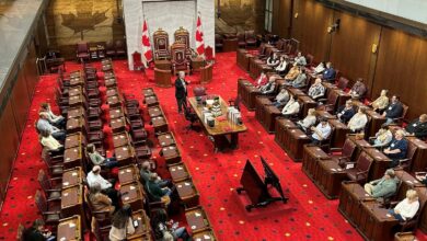Legislative chamber with red carpet, rows of desks, and a central dais flanked by Canadian flags and gilded chairs under a marble backdrop.