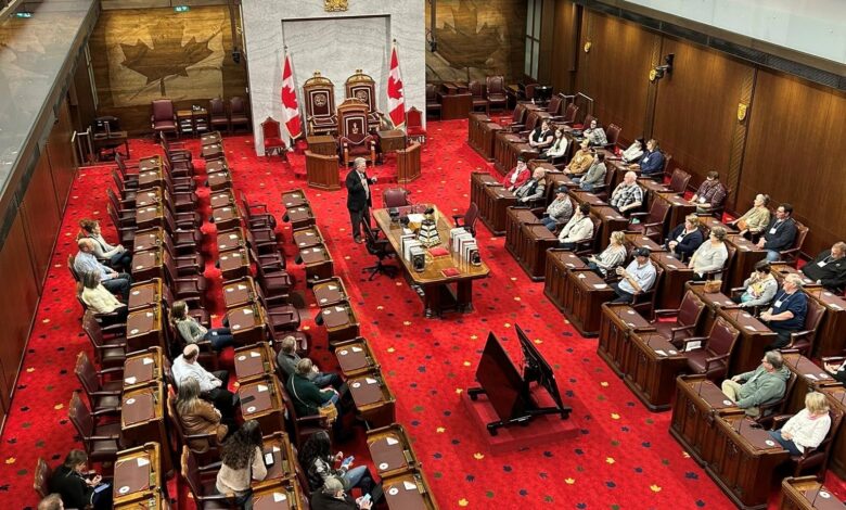 Legislative chamber with red carpet, rows of desks, and a central dais flanked by Canadian flags and gilded chairs under a marble backdrop.