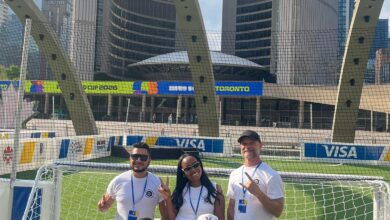Five staff members pose for a group photo in white shirts on a small soccer goal, in a city stadium with VISA banners and tall buildings behind them.