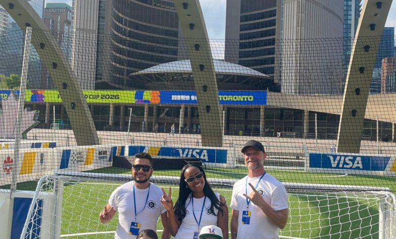 Five staff members pose for a group photo in white shirts on a small soccer goal, in a city stadium with VISA banners and tall buildings behind them.