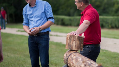 Two men outdoors laughing as one watches a man in a red shirt handle a large log on a splitting/sawing setup by a grassy area.