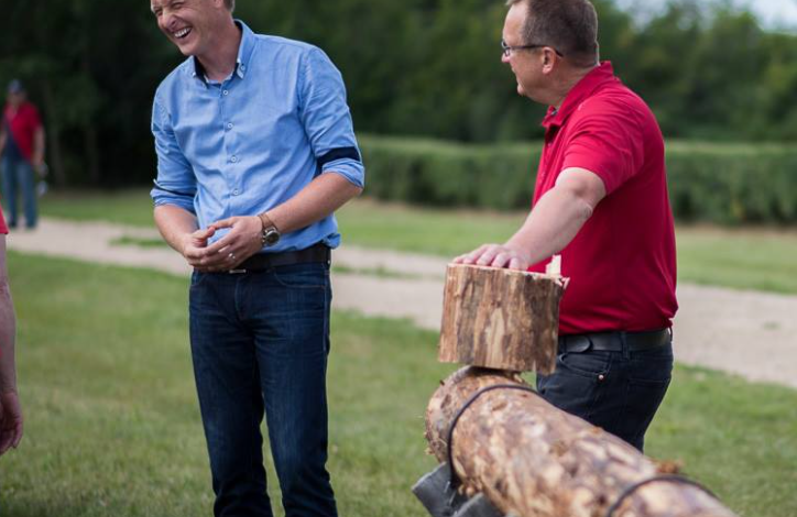 Two men outdoors laughing as one watches a man in a red shirt handle a large log on a splitting/sawing setup by a grassy area.