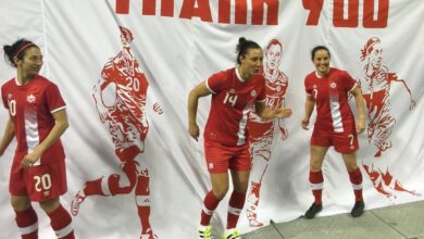 Three female soccer players in red uniforms stand in front of a white banner that says 'THANK YOU'.
