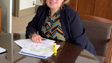 Woman in a dark blazer sits at a glass conference table, papers and a yellow highlighter in front of her in an office setting.