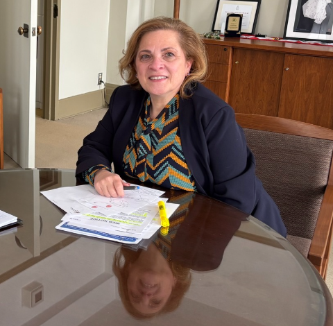 Woman in a dark blazer sits at a glass conference table, papers and a yellow highlighter in front of her in an office setting.