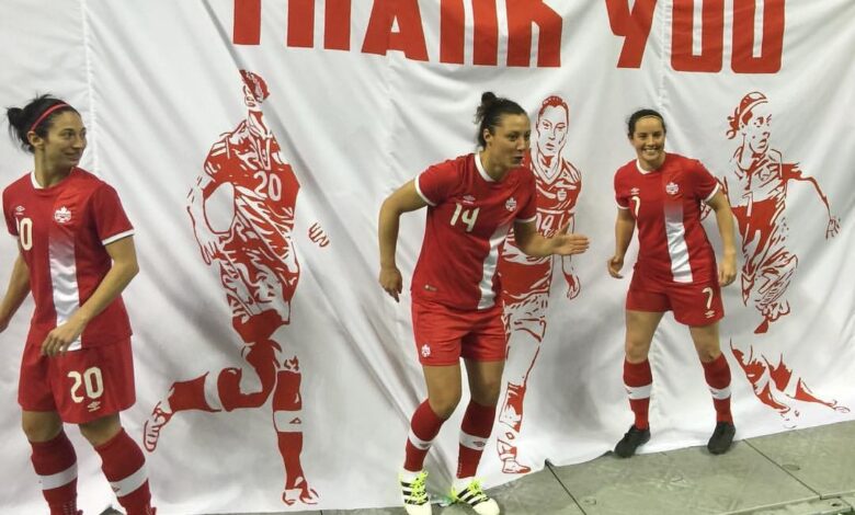 Three female soccer players in red uniforms stand in front of a white banner that says 'THANK YOU'.