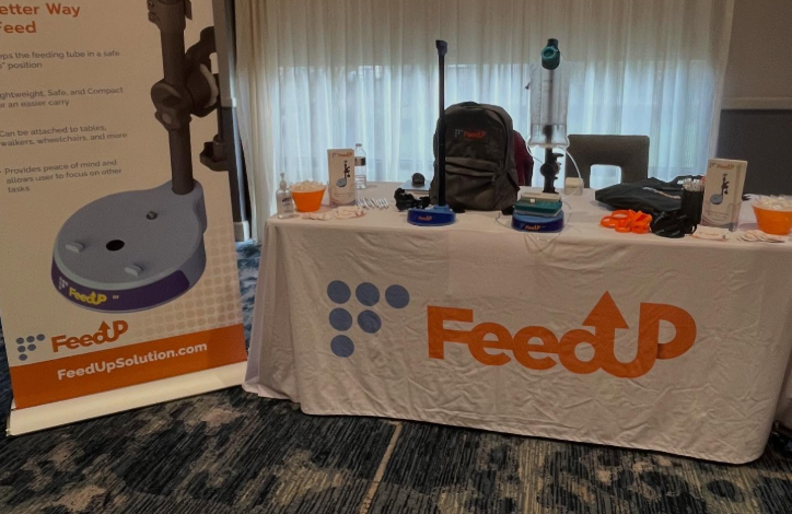 Table setup for FeedUp at a conference, featuring a branded banner and a table with medical feeding equipment and packets.