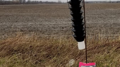 Soil sampling device with a black helical auger attached to a pole and a labeled tag in a bare farmland field.