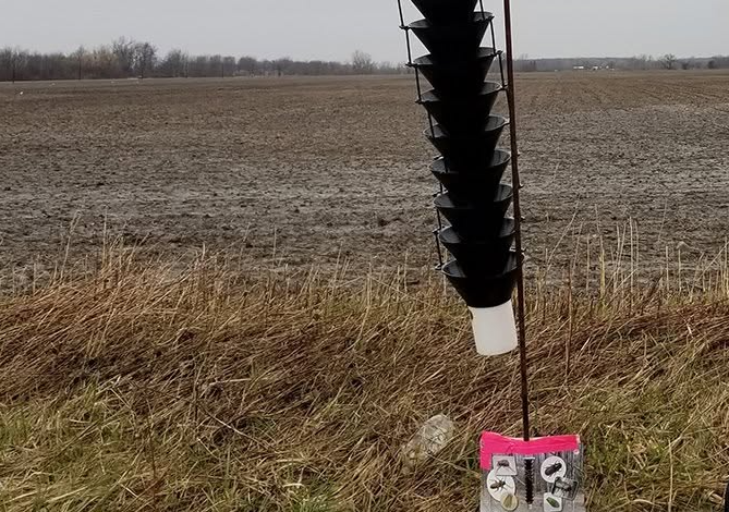Soil sampling device with a black helical auger attached to a pole and a labeled tag in a bare farmland field.