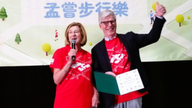 Two presenters on stage in red shirts; woman speaks into a microphone, man holds a certificate and raises his fist.